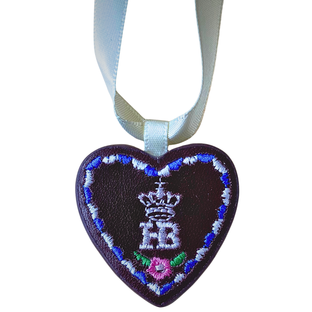 Herzförmiger Anhänger aus braunem Leder mit „HB“-Schriftzug, befestigt an einem silbrig-weißen Satinband.